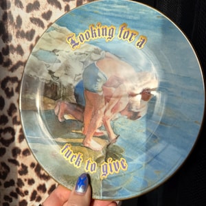 May include: A round ceramic decorative plate featuring a blue and white illustration of a person kneeling by the water. The text "Looking for a fuck to give" is printed in gold lettering across the top and bottom of the plate, which has a gold rim.