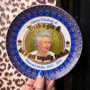 May include: A decorative ceramic plate featuring a portrait of Queen Elizabeth II. The border is deep blue with gold filigree. Text reads: Queen Elizabeth II, Grab a gin & let's get royally fucked, Diamond Jubilee 1952 - 2012, and the ER emblem.