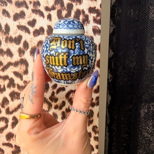 May include: A small, round, blue and white ceramic jar with a matching lid. The jar features a decorative floral pattern and the text "Don't sniff my hamster" written in a bold, black, gothic-style font across the front of the rounded body.