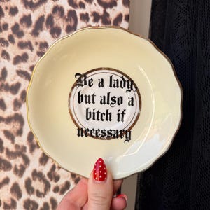 May include: A cream-coloured ceramic saucer with a scalloped edge and a gold-toned rim. The centre features black text in a gothic-style font that reads: Be a lady, but also a bitch if necessary. The saucer has a smooth, glossy finish.