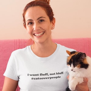 Puede incluir: Una mujer con una camiseta blanca con el texto "I want fluff, not bluff #catsoverpeople" está sentada en un sofá rosa, sosteniendo dos gatos. Uno es gris y blanco, el otro es calicó.