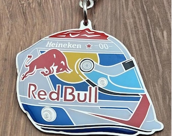 Könnte beinhalten: Ein Metallschlüsselanhänger in Form eines Rennhelms mit einer roten Stiergrafik, dem Schriftzug Red Bull und dem Text Heineken 00 mit einem roten Stern. Das Design umfasst blaue, gelbe, weiße und rote Farben mit einem silberfarbenen Metallring.