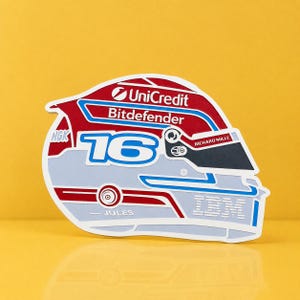 Op de afbeelding: Een sticker in de vorm van een racehelm, met het nummer 16 in wit en blauw. Het helmontwerp bevat de logo's UniCredit en Bitdefender, samen met de namen "JULES" en "RICHARD MILLE". De helm is voornamelijk rood, wit en blauw.
