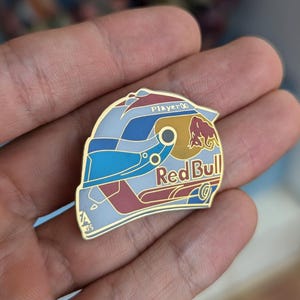 K&ouml;nnte beinhalten: Ein Metall-Emaille-Pin in Form eines Rennhelms mit einem blau-wei&szlig;-roten Farbschema. Das Design enth&auml;lt das Red Bull-Logo, den Text Player 00 und ein EA Sports-Logo. Der Pin hat goldfarbene Metallumrandungen und feine Details.