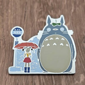 Puede incluir: Un pin metálico con un personaje azul de Totoro y una niña con un paraguas rojo. El pin incluye texto japonés y una señal de parada de autobús. El fondo es azul claro con un contorno plateado.