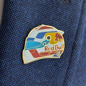 K&ouml;nnte beinhalten: Eine Metall-Emaille-Anstecknadel in Form eines Rennhelms mit einem Farbschema in Hellblau, Dunkelblau und Rot. Das Design enth&auml;lt das rote und gelbe Red Bull-Logo, den Schriftzug Player und ein kleines EA Sports-Logo unten links.