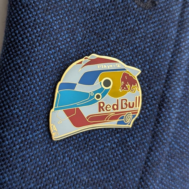 K&ouml;nnte beinhalten: Eine Metall-Emaille-Anstecknadel in Form eines Rennhelms mit einem Farbschema in Hellblau, Dunkelblau und Rot. Das Design enth&auml;lt das rote und gelbe Red Bull-Logo, den Schriftzug Player und ein kleines EA Sports-Logo unten links.