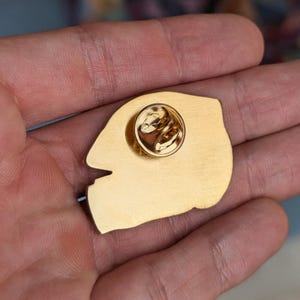 K&ouml;nnte beinhalten: Eine Anstecknadel aus goldfarbenem Metall mit einer glatten, geb&uuml;rsteten Oberfl&auml;che auf der R&uuml;ckseite. Die R&uuml;ckseite der Nadel zeigt einen runden Verschlussmechanismus mit einem zentralen Stift zur sicheren Befestigung an Stoffen.