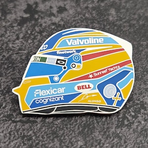 Pode incluir: Um autocolante colorido em forma de capacete de corrida. O capacete apresenta detalhes em azul, amarelo, vermelho e branco, com os logótipos "Valvoline", "Flexicar cognizant" e "Sunreef Yachts". O nome "ALONSO" também é visível.