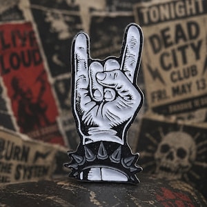 May include: A black and white enamel pin in the shape of a hand making the rock and roll hand gesture. The pin has a black base with silver spikes and a white hand with black outlines. The background features vintage concert posters.