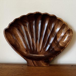 May include: A wooden seashell-shaped bowl with a rich, dark brown finish. The bowl features a scalloped edge and carved vertical grooves, creating a textured surface. The bowl is designed with a flat base for stability and is suitable for decorative or functional use.