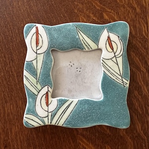 Könnte beinhalten: Ein quadratischer Keramik-Bilderrahmen mit türkisfarbenem Rand und floralem Design. Der Rahmen zeigt weiße Calla-Lilien mit orangefarbenen Zentren und grünen Blättern. Der Rahmen ist etwa 13 cm groß.