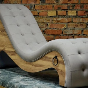 Könnte beinhalten: Eine moderne Chaiselongue mit geschwungenem Design. Sitzfläche und Rückenlehne sind mit hellgrauem Stoff mit Knopfheftung gepolstert. Der Rahmen ist aus Holz mit natürlicher Oberfläche und hat schwarze Metallringgriffe.