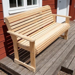 Könnte beinhalten: Eine hellfarbene Holzbank mit Lattenrücken und -sitzfläche sowie Armlehnen. Die Bank ist aus Naturholz gefertigt und für den Außenbereich konzipiert. Die Bank ist etwa 152 cm lang.