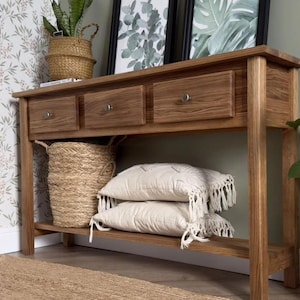 May include: A wooden console table with three drawers and a lower shelf. The table is a light brown color and has a natural wood grain. Two white pillows with fringe are on the shelf, along with a woven basket.