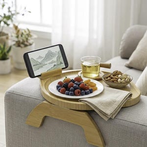May include: A bamboo sofa arm tray with a built-in phone holder. The tray holds a plate of berries, a bowl of nuts, a glass of tea, and a linen napkin. A smartphone displays a landscape image.