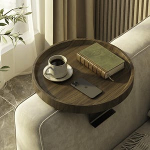 May include: A round, wooden side table with a dark finish, holding a cup of coffee, a book, and a smartphone. The table is placed on the arm of a beige sofa, creating a cozy and functional space for relaxation and convenience.
