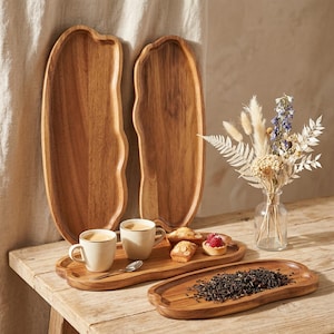 May include: Wooden serving trays in various sizes, showcasing a natural wood grain. Two large, organically shaped trays stand behind a smaller tray holding coffee cups and pastries. A vase of dried flowers and a tray of tea leaves complete the display.