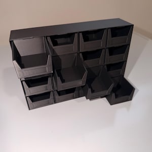 May include: A black, wall-mounted storage organizer with sixteen individual compartments. Each compartment is open-faced and angled for easy access. The organizer is made of a matte black material and is designed for small item storage.