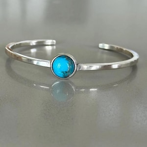May include: A silver cuff bracelet featuring a single, round, bright blue turquoise stone set in the centre. The metal band has a smooth, polished finish and an open design, allowing it to be slipped onto the wrist with ease and comfort.