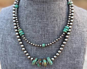 Puede incluir: Un collar de dos hileras con cuentas plateadas pulidas intercaladas con piedras redondas de color turquesa. La hilera más corta incluye cuentas turquesas individuales, mientras que la más larga presenta un grupo de cuatro cuentas turquesas.