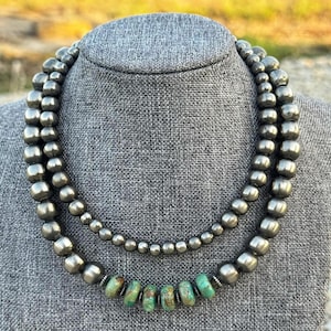May include: A double-strand necklace featuring polished metallic silver-toned beads of varying sizes. The inner strand consists of smaller uniform beads, while the outer strand features larger beads with six green and brown turquoise-style stones.