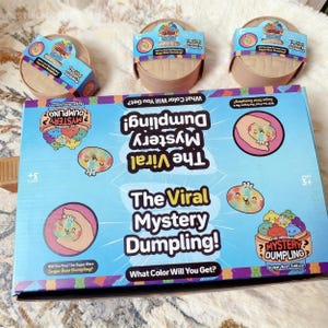 Puede incluir: Una caja azul claro y tres recipientes redondos con el texto "The Viral Mystery Dumpling!" y "Sugar Bear Edition." La caja presenta ilustraciones de dibujos animados de albóndigas y el texto "What Color Will You Get?" y "Will You Find The Super Rare Sugar Bear Dumpling?"