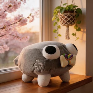 Könnte beinhalten: Ein graues Plüschtier mit großen, runden Augen und einer bunten Fliege. Das Stofftier hat ein vierbeiniges Design und ein dekoratives Emblem an der Seite. Es sitzt auf einer Holzoberfläche in der Nähe eines Fensters mit einer Hängepflanze.