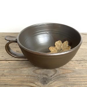 May include: A dark brown ceramic bowl with a handle and a decorative flower inside. The bowl is set on a wooden surface. The handle is curved and attached to the side of the bowl.