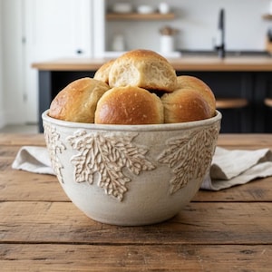 May include: A round, off-white ceramic bowl featuring a raised, textured maple leaf pattern around the exterior, filled with a mound of golden-brown, baked dinner rolls. The bowl has a matte finish and sits on a wooden surface.