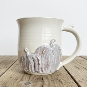 May include: A cream-colored ceramic mug featuring two raised, sculpted pumpkin designs on the front. The pumpkins are finished in a light, muted lavender-grey glaze with visible texture and ridges, contrasting against the smooth, glossy off-white body of the mug.