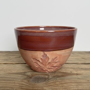 May include: A handmade ceramic bowl with a two-tone design. The upper portion is a glossy, deep reddish-brown, while the lower half is a lighter, matte terracotta color. A detailed leaf design is carved into the terracotta section. The bowl sits on a wooden surface.