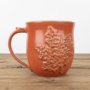May include: A burnt orange ceramic mug featuring a prominent, raised relief of a maple leaf on its side. The mug has a smooth, glossy finish on the body and a textured, detailed surface on the leaf design, complete with a curved handle on the left.