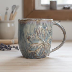 May include: A handmade ceramic mug with a large, embossed flower design. The mug features a blue and tan glaze with a unique handle. The mug is sitting on a wooden surface.