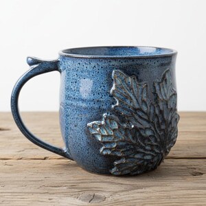 May include: A ceramic mug with a speckled blue glaze featuring a prominent, raised relief of a large maple leaf on the side. The mug has a curved handle and a smooth, rounded body, showcasing a deep blue color with darker blue speckles.