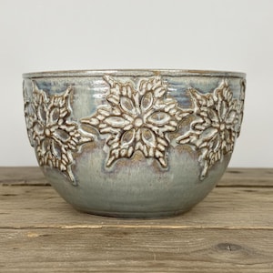 May include: A ceramic bowl with a light blue and grey glazed finish, featuring a repeating embossed snowflake pattern around the exterior. The bowl has a rounded shape and a smooth, glossy texture, showcasing a blend of cool tones.