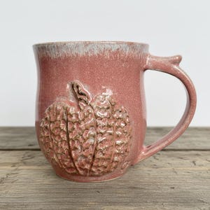 May include: A ceramic mug with a dusty rose glaze and a white rim. The exterior features a raised, textured pumpkin relief design with a brown wash in the crevices. The mug has a curved handle and a rounded body, showcasing a handcrafted finish.