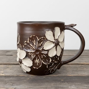 May include: A brown ceramic mug with white floral designs. The flowers are raised and textured, adding dimension to the mug's surface. The handle has a leaf-like detail. The mug sits on a wooden surface.