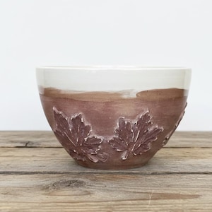 May include: A ceramic bowl featuring a two-tone design with a smooth white upper section and a textured brown lower section. The exterior is decorated with embossed maple leaf patterns and a small heart shape integrated into the clay surface.