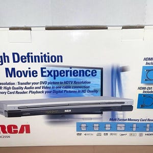 May include: A white cardboard box for an RCA DRC255N DVD player. The box features the text High Definition Movie Experience, an image of the silver and black player, and icons indicating an HDMI cable and HDMI-DVI cable are included inside.