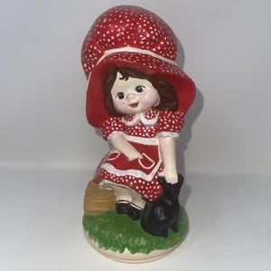 May include: A ceramic figurine of a girl in a red dress with white polka dots and a matching hat. The girl is holding a black cat and sitting on a patch of green grass. The figurine has a vintage, handcrafted appearance.