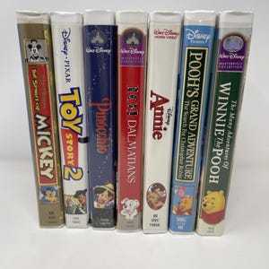 May include: A collection of seven Walt Disney VHS tapes standing upright, including The Spirit of Mickey, Toy Story 2, Pinocchio, 101 Dalmatians, Annie, Pooh's Grand Adventure: The Search for Christopher Robin, and The Many Adventures of Winnie the Pooh.