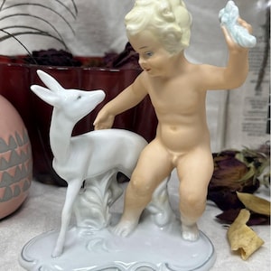 Może przedstawiać: Porcelanowa figurka przedstawiająca postać cherubina i białego jelenia. Cherubin, z blond włosami i niebieskimi oczami, trzyma jasnoniebieski przedmiot. Jeleń stoi obok cherubina na podstawie ze srebrnym obramowaniem.