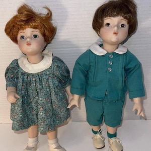 May include: Two porcelain dolls, a girl and a boy, stand side-by-side. The girl doll wears a teal dress with a floral pattern and white collar. The boy doll is dressed in a teal jacket and shorts with a white collar. Both dolls have painted faces and wear shoes.