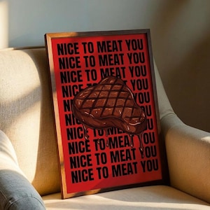 Op de afbeelding: Een ingelijste print met een rode achtergrond en de zin "NICE TO MEAT YOU" herhaald in zwart. Een cartoon steak illustratie is gecentreerd op de print. De print wordt weergegeven op een beige fauteuil.