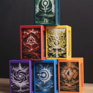 May include: A stack of six colorful, rectangular boxes with unique, detailed illustrations. Each box has a different color frame: green, red, yellow, purple, blue, and orange. The illustrations feature abstract designs in white and other colors.