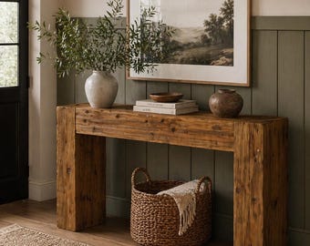 Puede incluir: Una consola de madera rústica con una impresión de paisaje enmarcada, una cesta tejida y jarrones decorativos. La mesa está hecha de madera gruesa y tosca. Una alfombra beige y paneles de pared verde oliva completan el diseño.