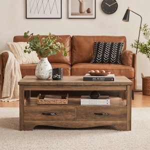 May include: A rectangular, wooden coffee table with two drawers and a lower shelf. The table is a medium brown color and has a distressed finish. A leather sofa and decorative items are in the background.