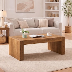 May include: A rectangular, solid wood coffee table with a natural finish. The table is in front of a light-colored sofa with decorative pillows. A rug and a lamp are also visible.