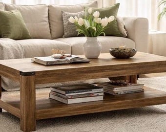 May include: A rectangular wooden coffee table with a lower shelf, holding several books. The table is in front of a beige sofa with green and patterned pillows. A white vase with white flowers and a decorative bowl are on the table.